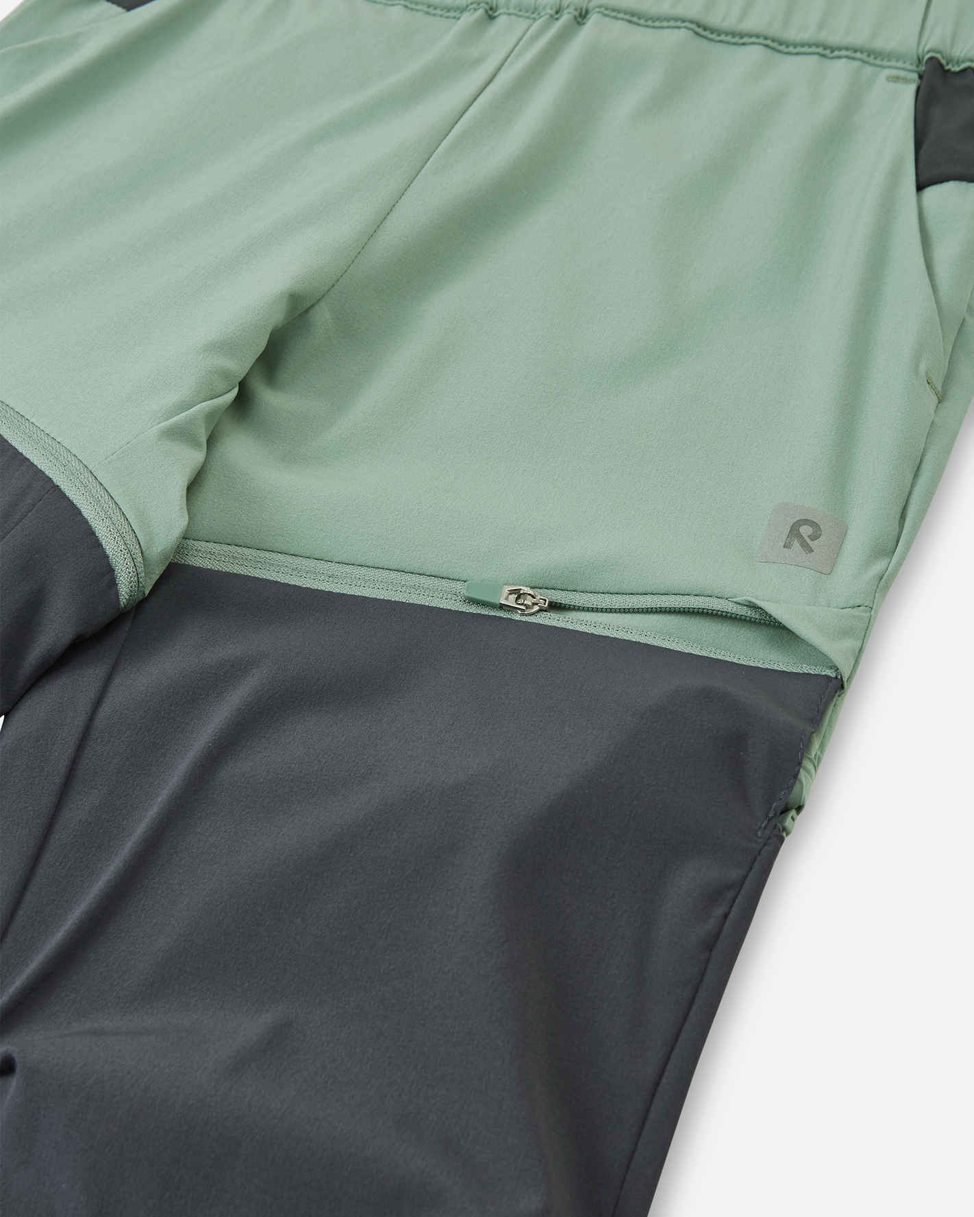 Macro close-up of the hidden zipper detail on the Reima Kaveris kids' stone green convertible pants, showing the durable hardware used to easily detach the dark grey lower leg panels.