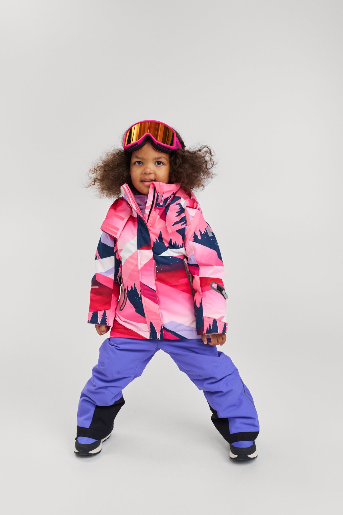 Studio lifestyle portrait of a young girl playfully posing in the soft coral mountain print Reima Kiiruna girls' ski jacket, styled with purple snow pants and pink ski goggles, demonstrating the jacket's comfortable, active fit designed for winter sports and alpine adventures.