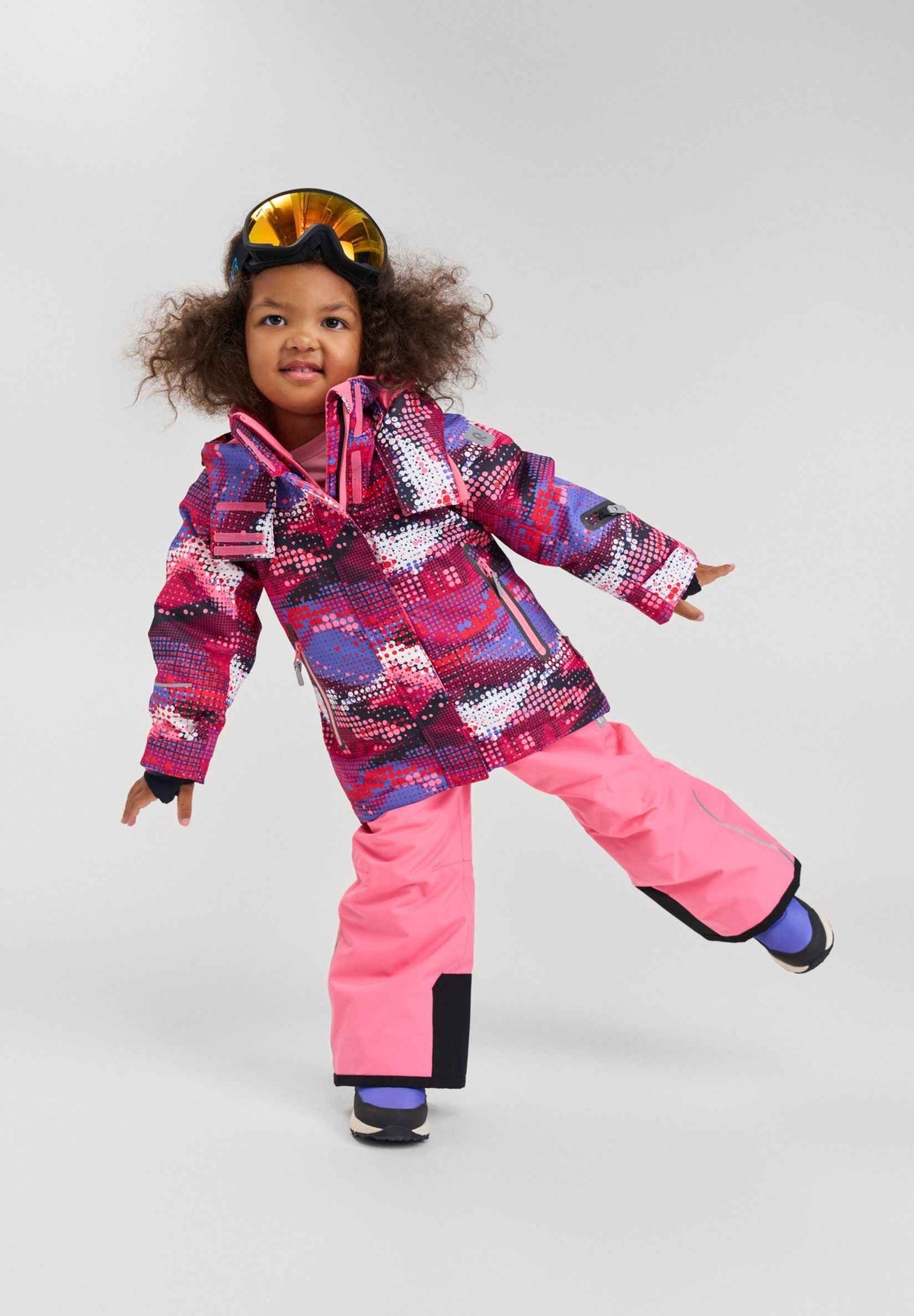 Studio lifestyle portrait of a young girl playfully posing in the rosy berry print Reima Kiiruna ski jacket paired with solid pink snow pants and ski goggles, showcasing the outfit's comfortable, active fit for winter sports.