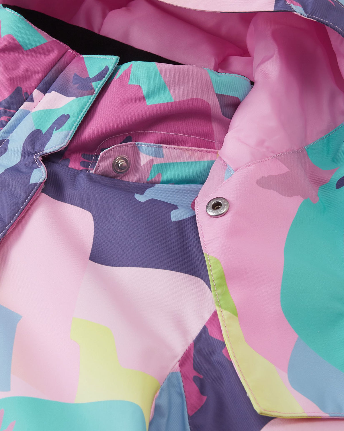 Macro detail shot of the collar on the Reima Kiiruna girls' ski jacket, highlighting the secure metallic snap buttons used to easily attach or remove the safety hood to the grey pink patterned fabric.