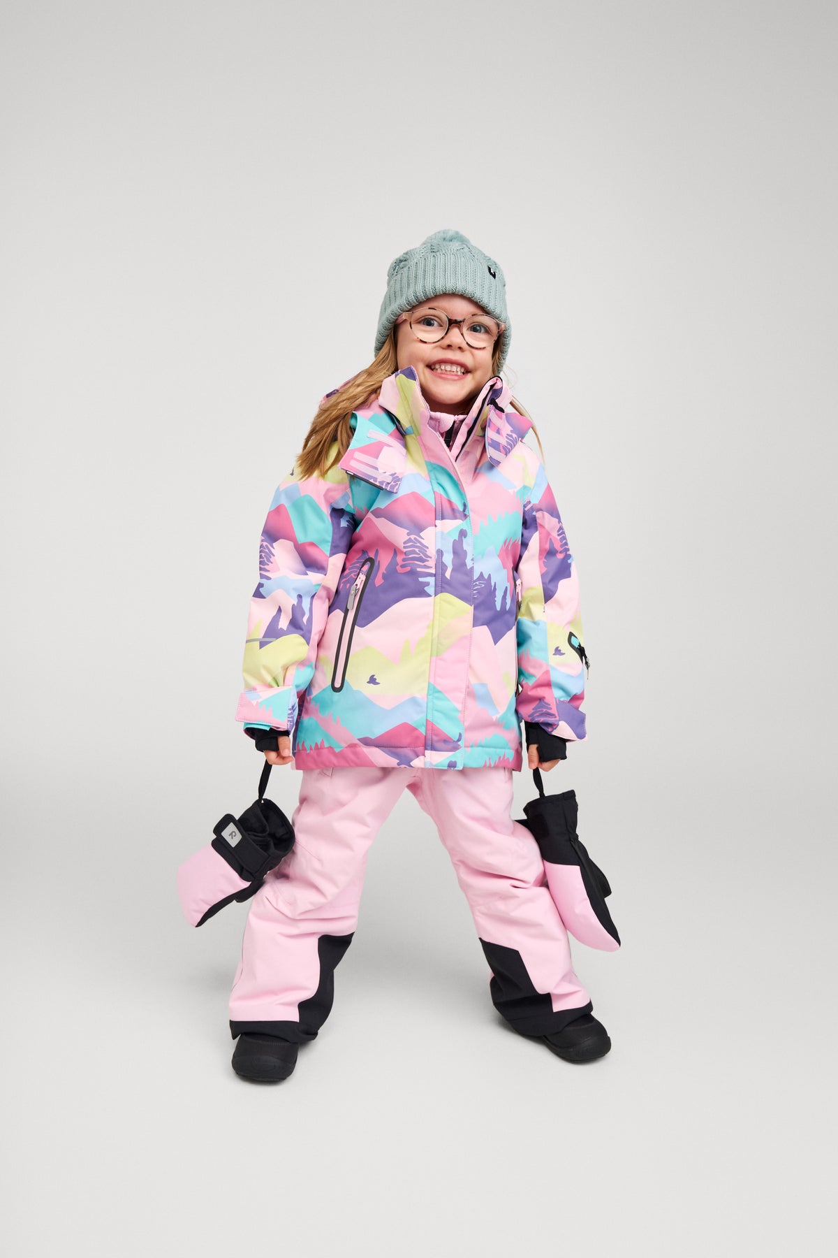 A young girl smiling and modeling the Reima Kiiruna waterproof girls' ski jacket in a vibrant grey, pink, and purple mountain print, fully outfitted for winter sports with matching pink snow pants, black winter mittens, and a knit beanie.