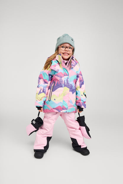 A young girl smiling and modeling the Reima Kiiruna waterproof girls' ski jacket in a vibrant grey, pink, and purple mountain print, fully outfitted for winter sports with matching pink snow pants, black winter mittens, and a knit beanie.