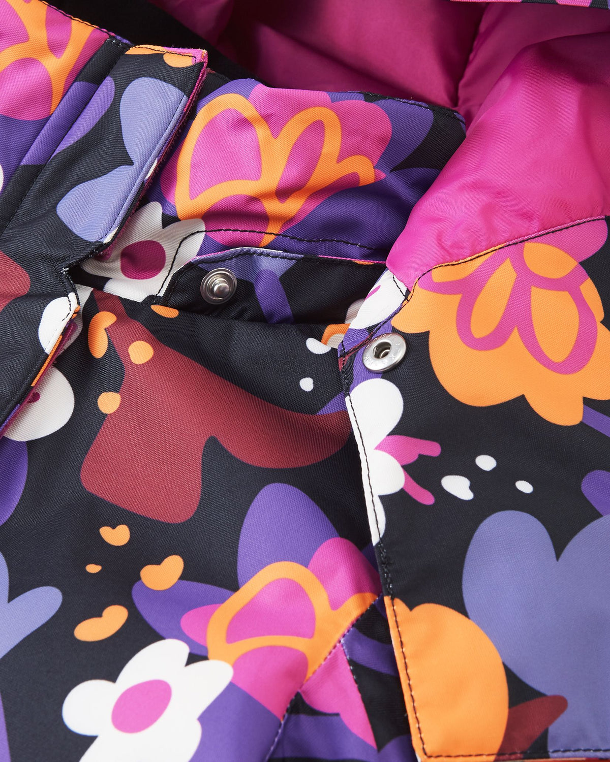 Macro detail shot of the collar on the Reima Kiiruna girls' ski jacket, highlighting the secure metallic snap buttons used to attach the removable safety hood to the black floral fabric.