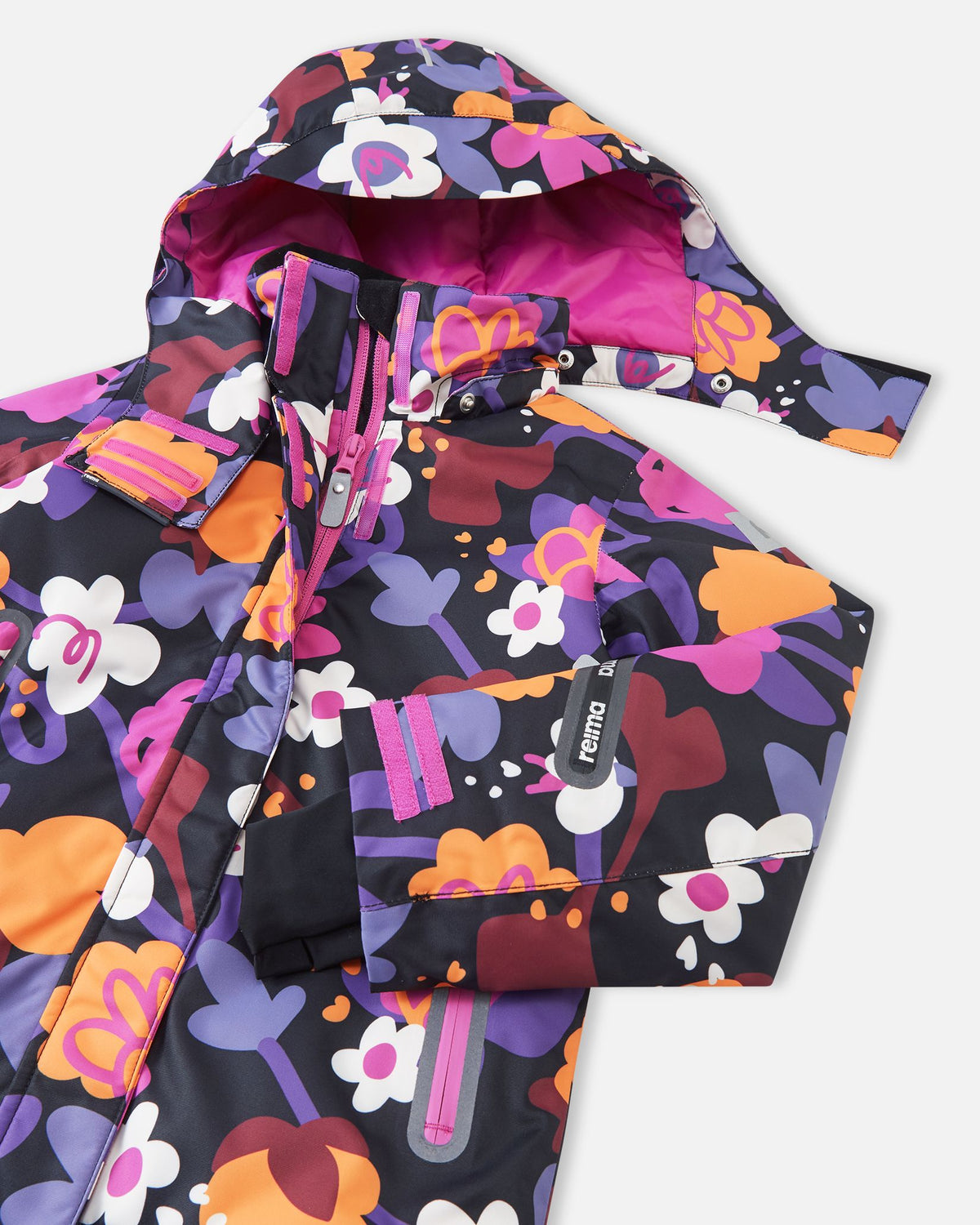 Close-up of the upper chest and collar on the Reima Kiiruna girls' ski jacket in black floral, detailing the snap buttons for the detachable safety hood, the protective front zipper, and the left sleeve ski pass pocket.