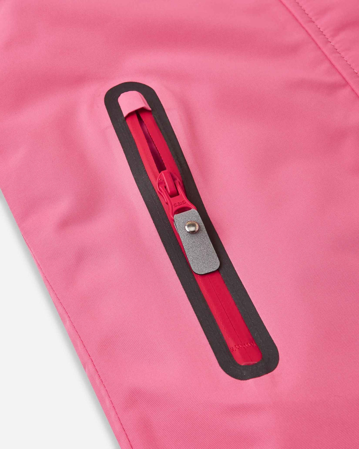 Close-up of the vertical zippered pocket on the solid soft coral Reima Kiiruna kids' snow jacket, featuring a water-resistant zipper track and a reflective pull tab for easy access with bulky ski gloves.