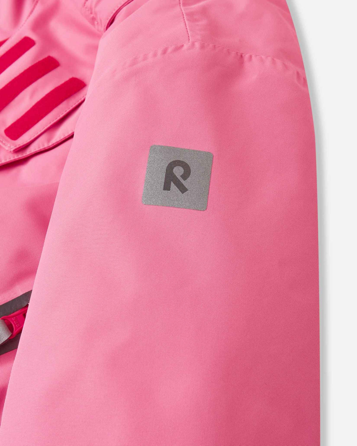Detail view of the solid pink sleeve on the Reima Kiiruna youth ski coat, highlighting the square reflective Reima logo patch designed for high visibility during dark winter afternoons.