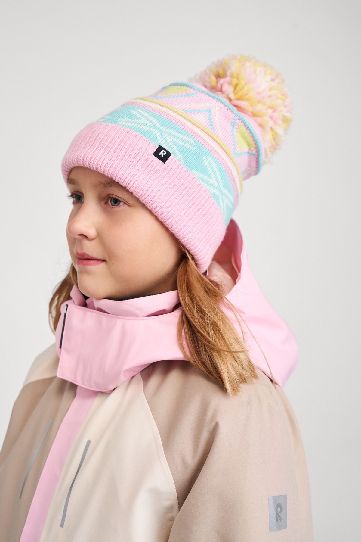 A young girl modeling the Reima Kirjoen light heather pink merino wool pom-pom winter hat paired with a winter parka, demonstrating fit, coverage, and cold-weather styling.