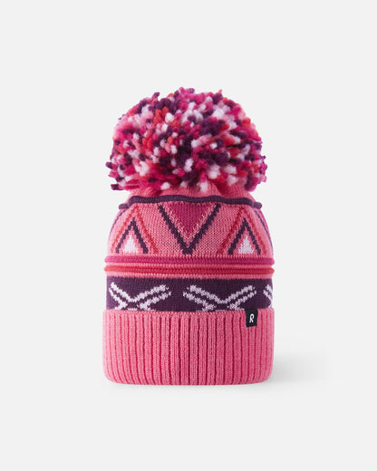 Front flat lay view of the soft coral pink Reima Kirjoen kids' merino wool beanie. The image highlights the colorful geometric jacquard knit pattern, a wide ribbed fold-up hem, and a matching multi-colored yarn pom-pom on top.