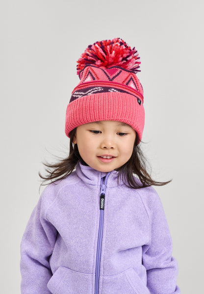 Lifestyle portrait of a young girl smiling while wearing the soft coral pink Reima Kirjoen kids' merino wool beanie paired with a light purple fleece jacket. The image demonstrates how the ribbed hem provides a secure, warm fit for optimal thermal control.