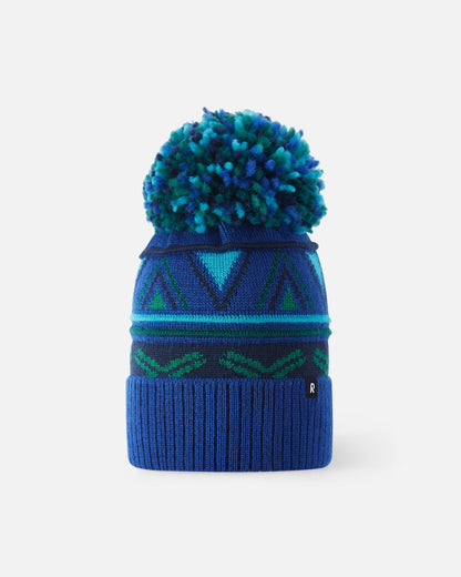 Front flat lay view of the twilight blue Reima Kirjoen kids' merino wool beanie. The image highlights the colorful geometric jacquard knit pattern with green and navy accents, a wide ribbed fold-up hem, and a matching multi-colored yarn pom-pom on top.