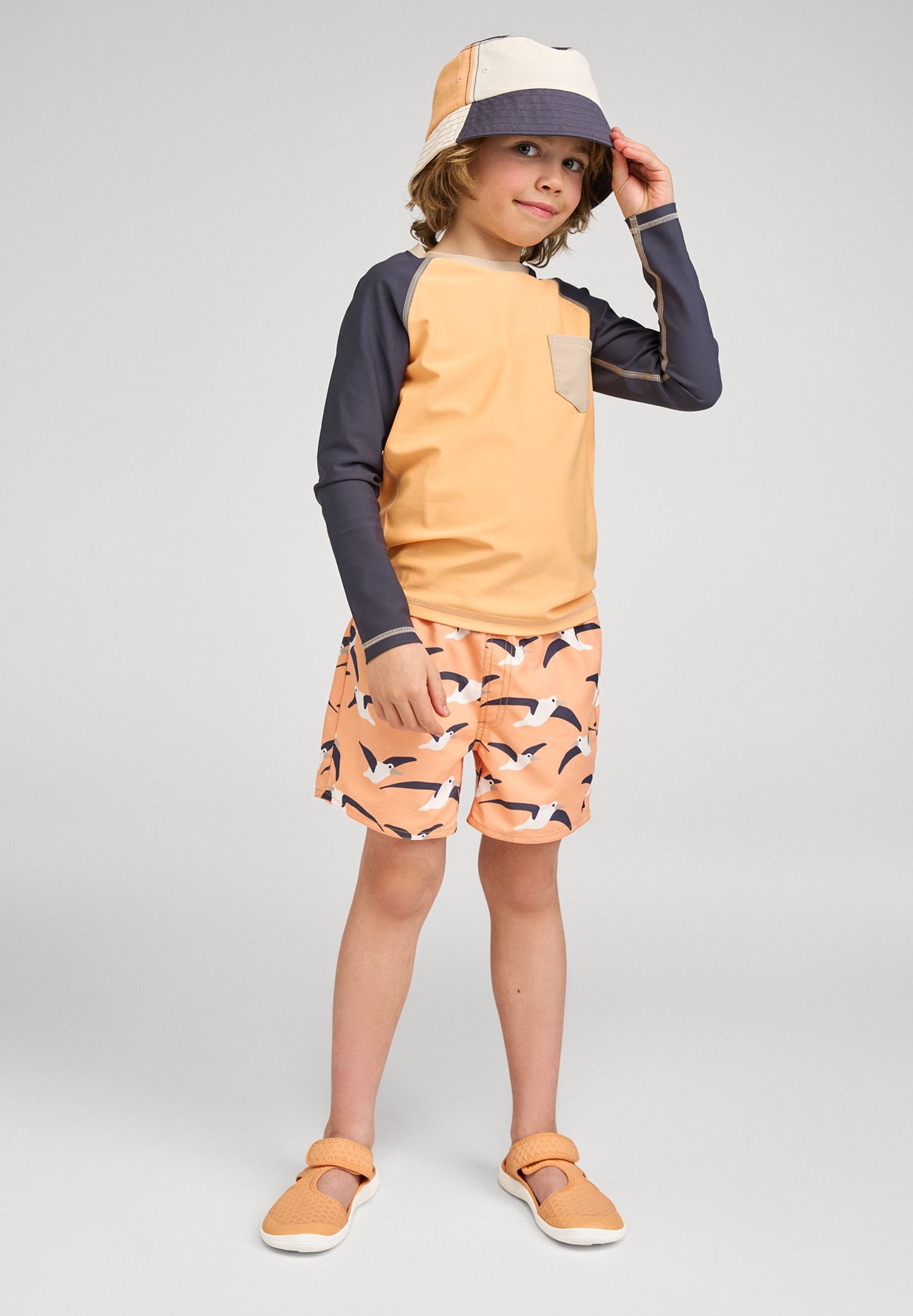 Lifestyle shot of a young boy wearing the Reima Kroolaus long-sleeve rash guard featuring UPF 50+ sun protection, styled with matching bird-print swim trunks, a bucket hat, and water sandals for summer beach play.