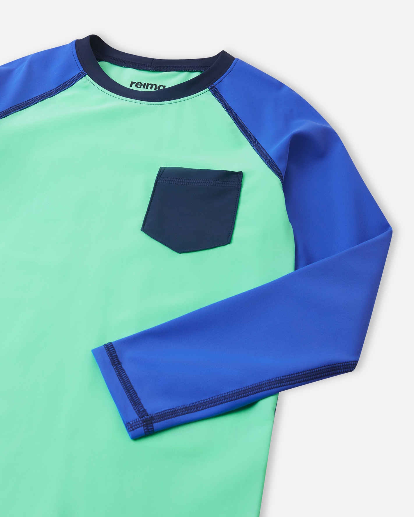 Close-up detail of the Reima Kroolaus kids' fresh mint sunproof rash guard, highlighting the contrasting blue chest pocket, flatlock seams to prevent chafing, and the Reima Sunproof logo on the inside collar.