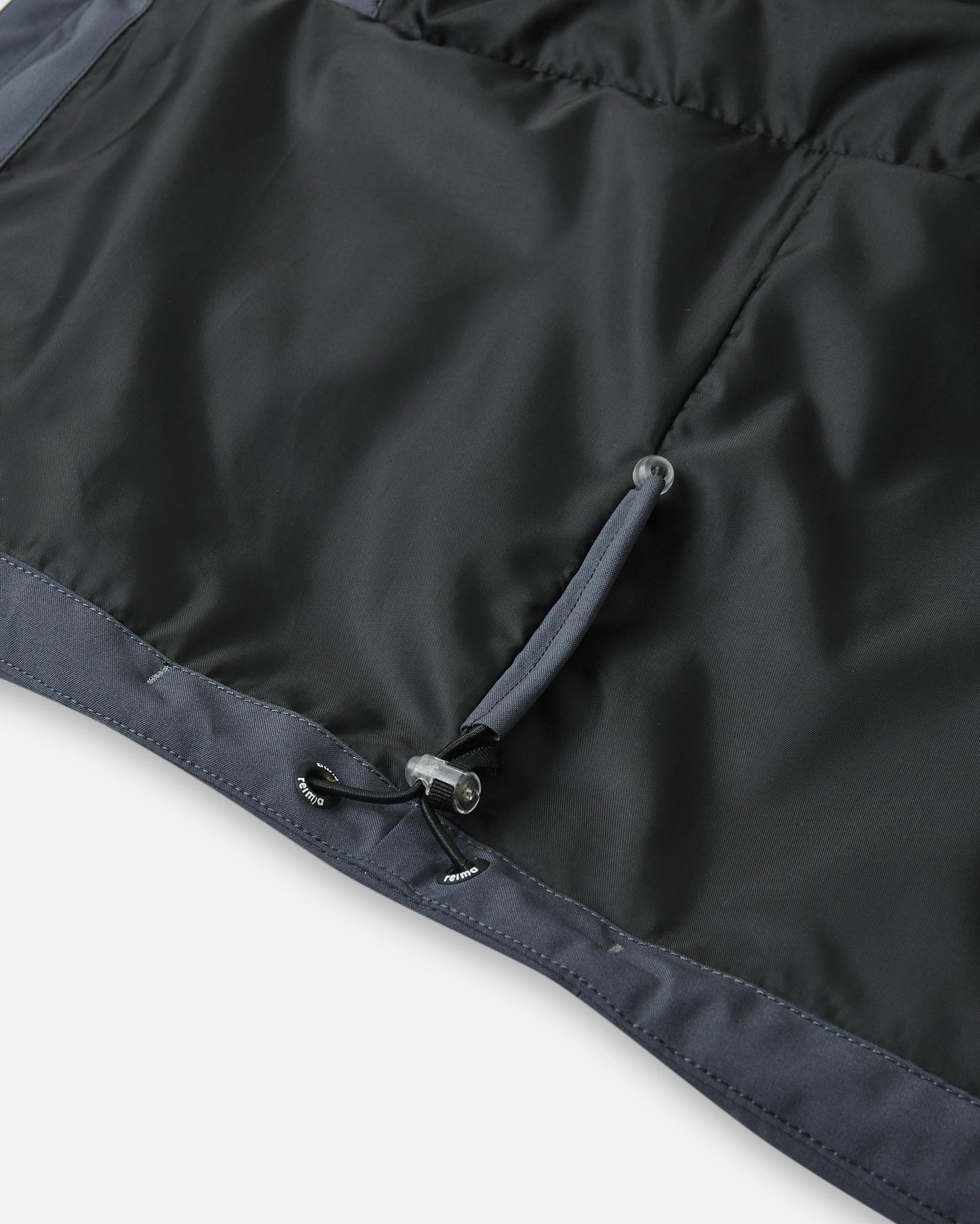 Close-up of the interior bottom hem of the Reima Kuosku kids ski jacket in Soft black, showing the adjustable elastic bungee cord and toggle system to customize the fit and block out wind.