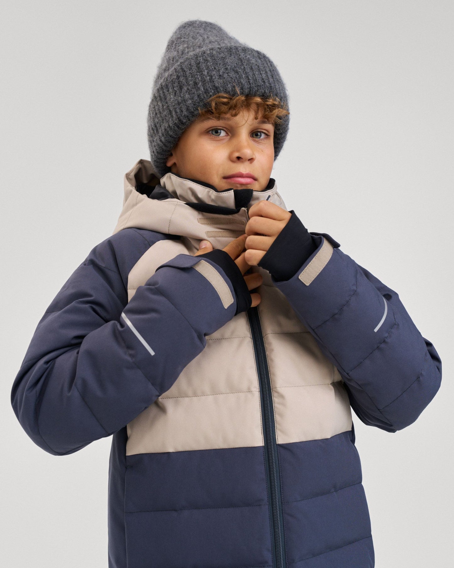 A young boy wearing a grey beanie and the color-blocked Reima Kuosku kids ski jacket in Soft black, pulling up the front collar zipper.