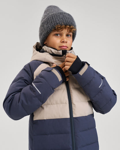 A young boy wearing a grey beanie and the color-blocked Reima Kuosku kids ski jacket in Soft black, pulling up the front collar zipper.