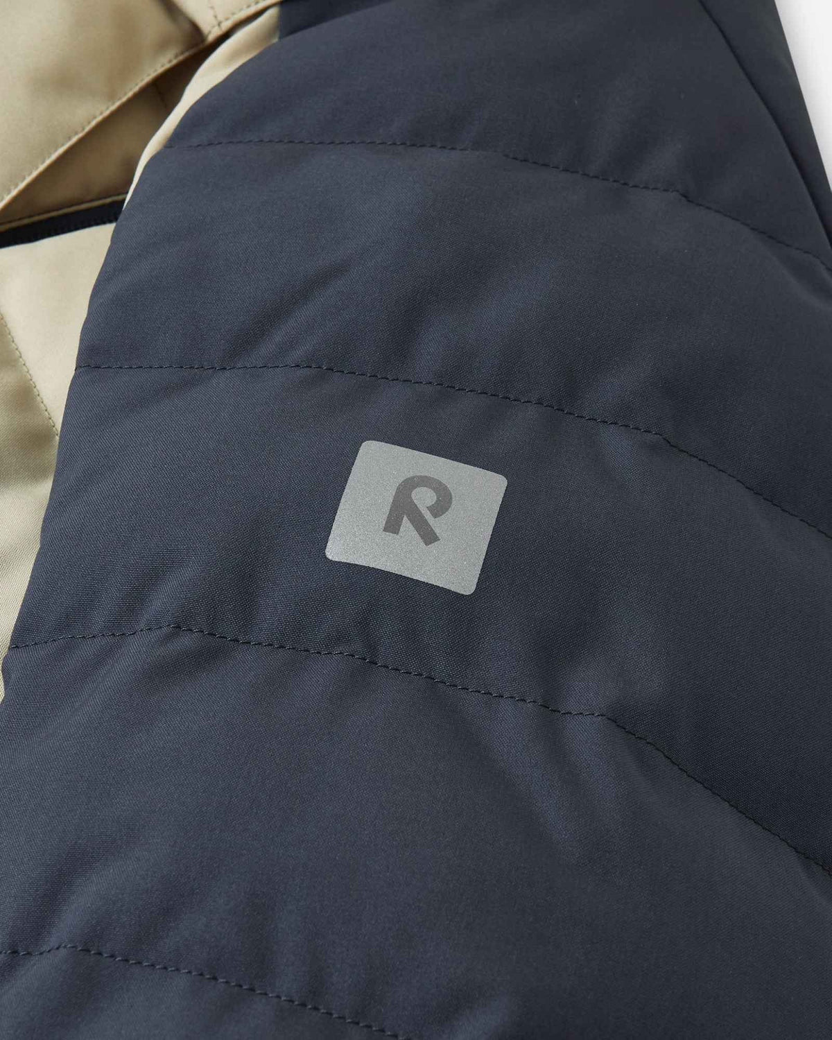 Close-up of the sleeve on the Reima Kuosku kids ski jacket in Soft black, displaying the reflective Reima 'R' logo patch designed for high visibility in low winter light.