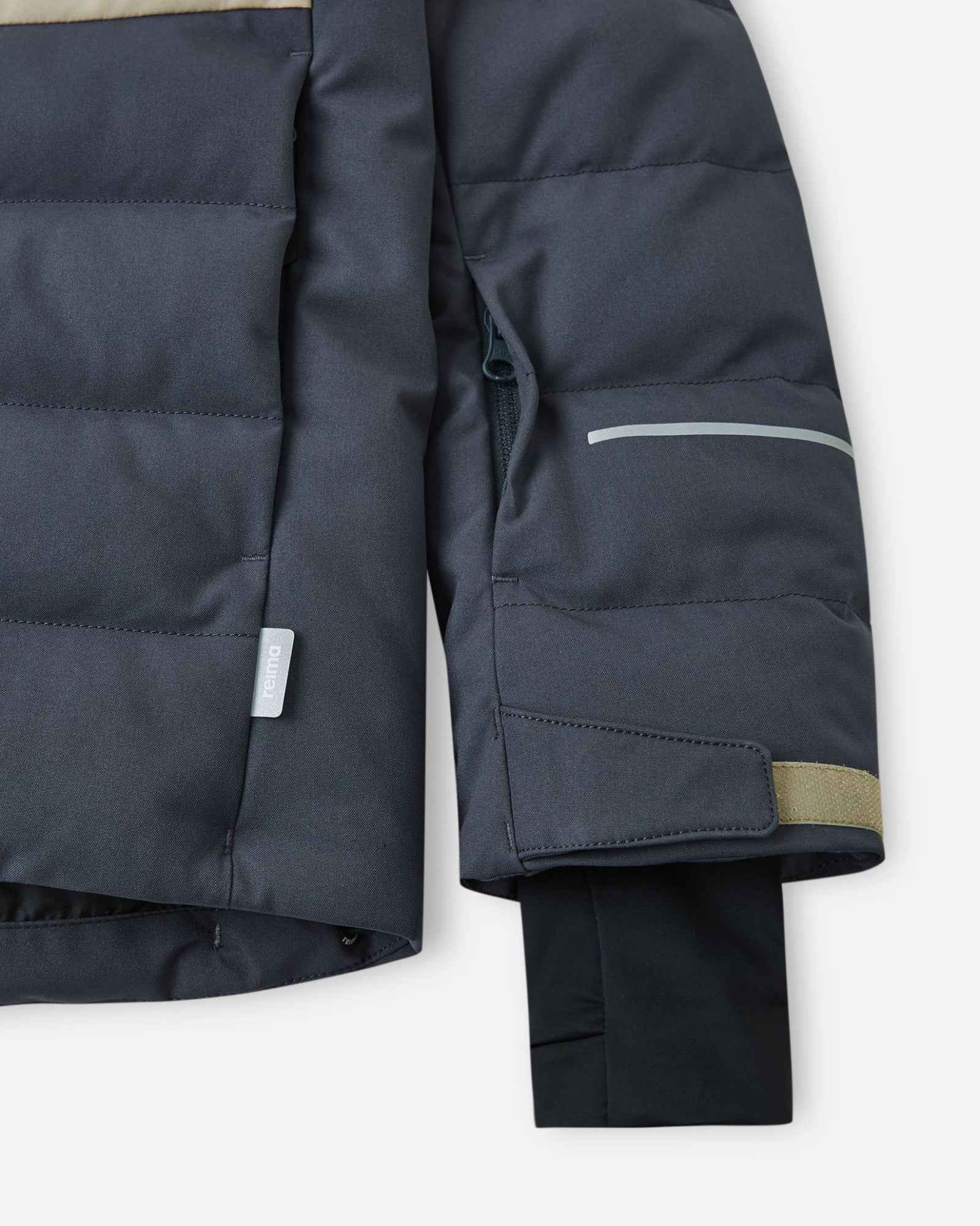 Close-up of the left sleeve on the Reima Kuosku kids ski jacket in Soft black, showing the zippered ski pass pocket, a reflective safety strip, and an adjustable cuff.