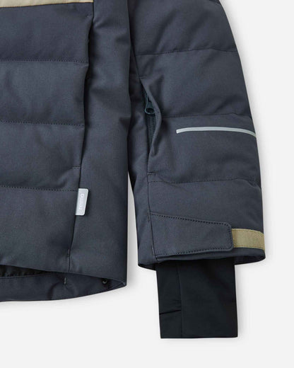 Close-up of the left sleeve on the Reima Kuosku kids ski jacket in Soft black, showing the zippered ski pass pocket, a reflective safety strip, and an adjustable cuff.