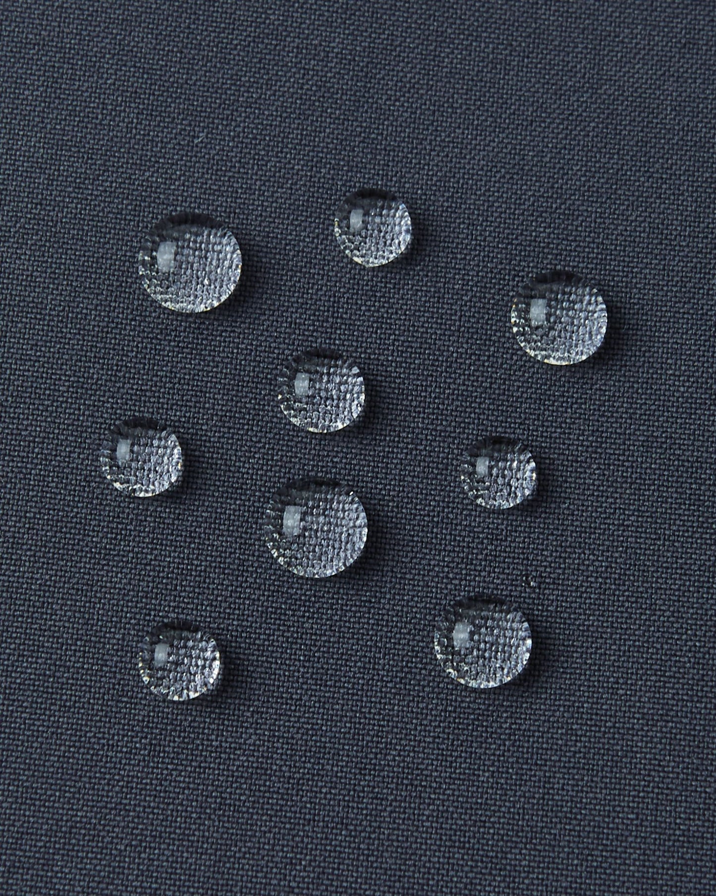A detailed close-up texture shot of several perfectly formed water droplets beading up cleanly on the dark surface of the water-repellent fabric of a Reima Kuosku kids ski jacket (Soft black color), demonstrating its durable, fluorocarbon-free performance.