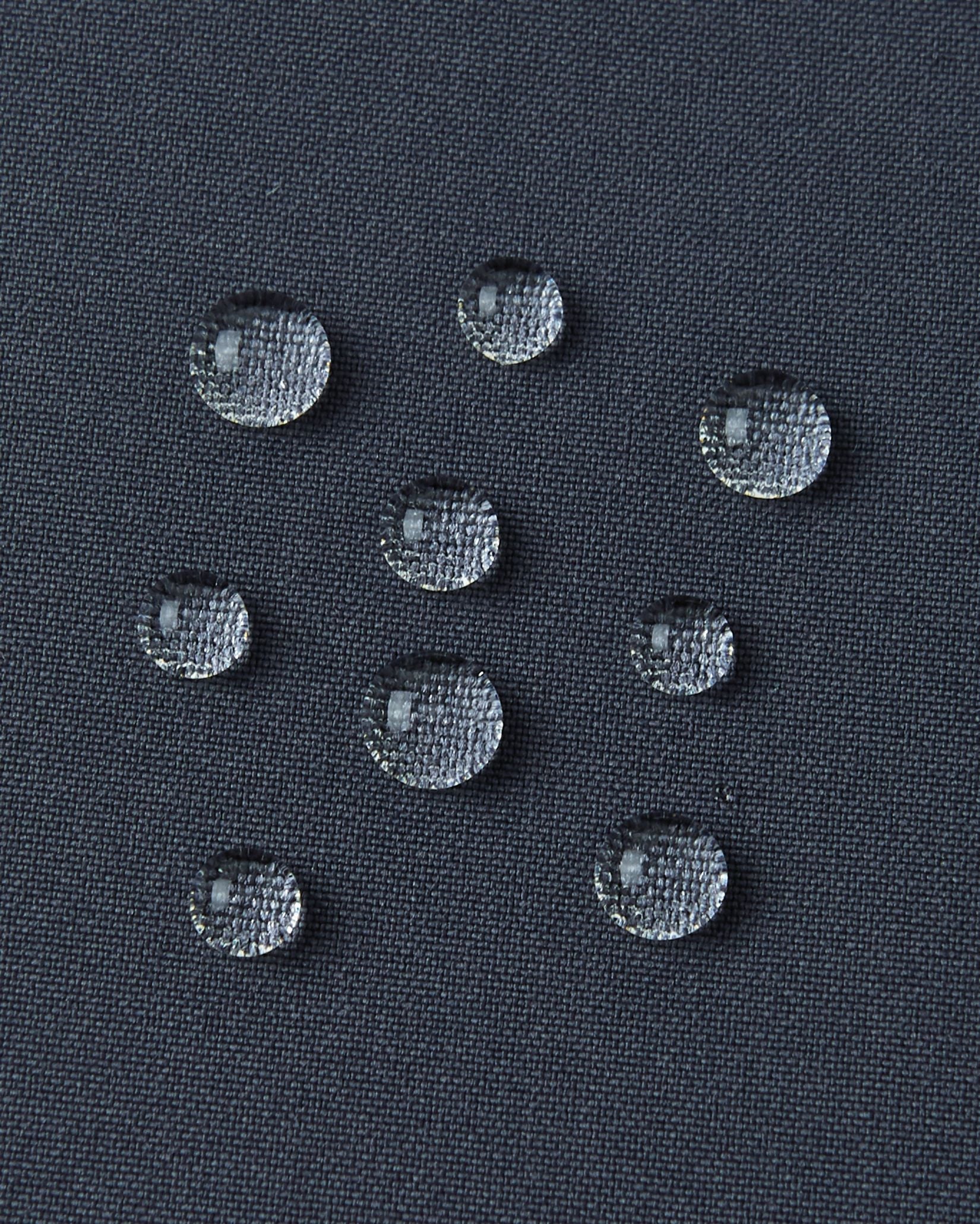 A detailed close-up texture shot of several perfectly formed water droplets beading up cleanly on the dark surface of the water-repellent fabric of a Reima Kuosku kids ski jacket (Soft black color), demonstrating its durable, fluorocarbon-free performance.