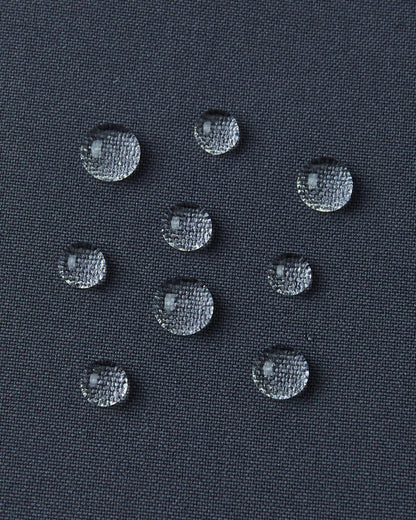 A detailed close-up texture shot of several perfectly formed water droplets beading up cleanly on the dark surface of the water-repellent fabric of a Reima Kuosku kids ski jacket (Soft black color), demonstrating its durable, fluorocarbon-free performance.