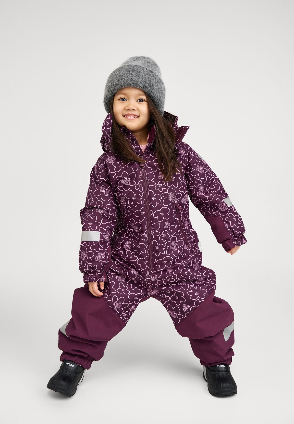 Lifestyle portrait of a smiling young girl modeling the deep purple Reima Kurikka kids' snowsuit with a matching floral top half, paired with a cozy grey knit beanie and winter boots.