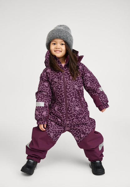 Lifestyle portrait of a smiling young girl modeling the deep purple Reima Kurikka kids' snowsuit with a matching floral top half, paired with a cozy grey knit beanie and winter boots.