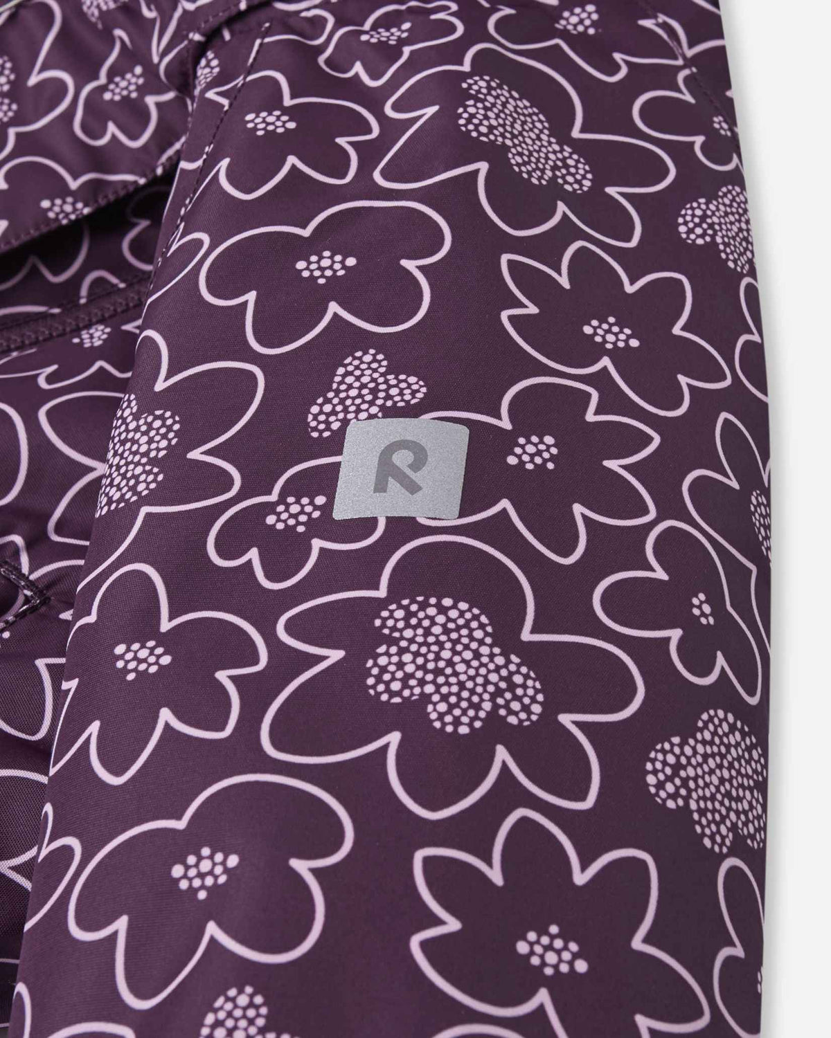 Detailed shot of the sleeve on the floral deep purple Reima Kurikka children's overall, focusing on the high-visibility reflective Reima badge integrated into the fabric to enhance child safety during dark winter afternoons.