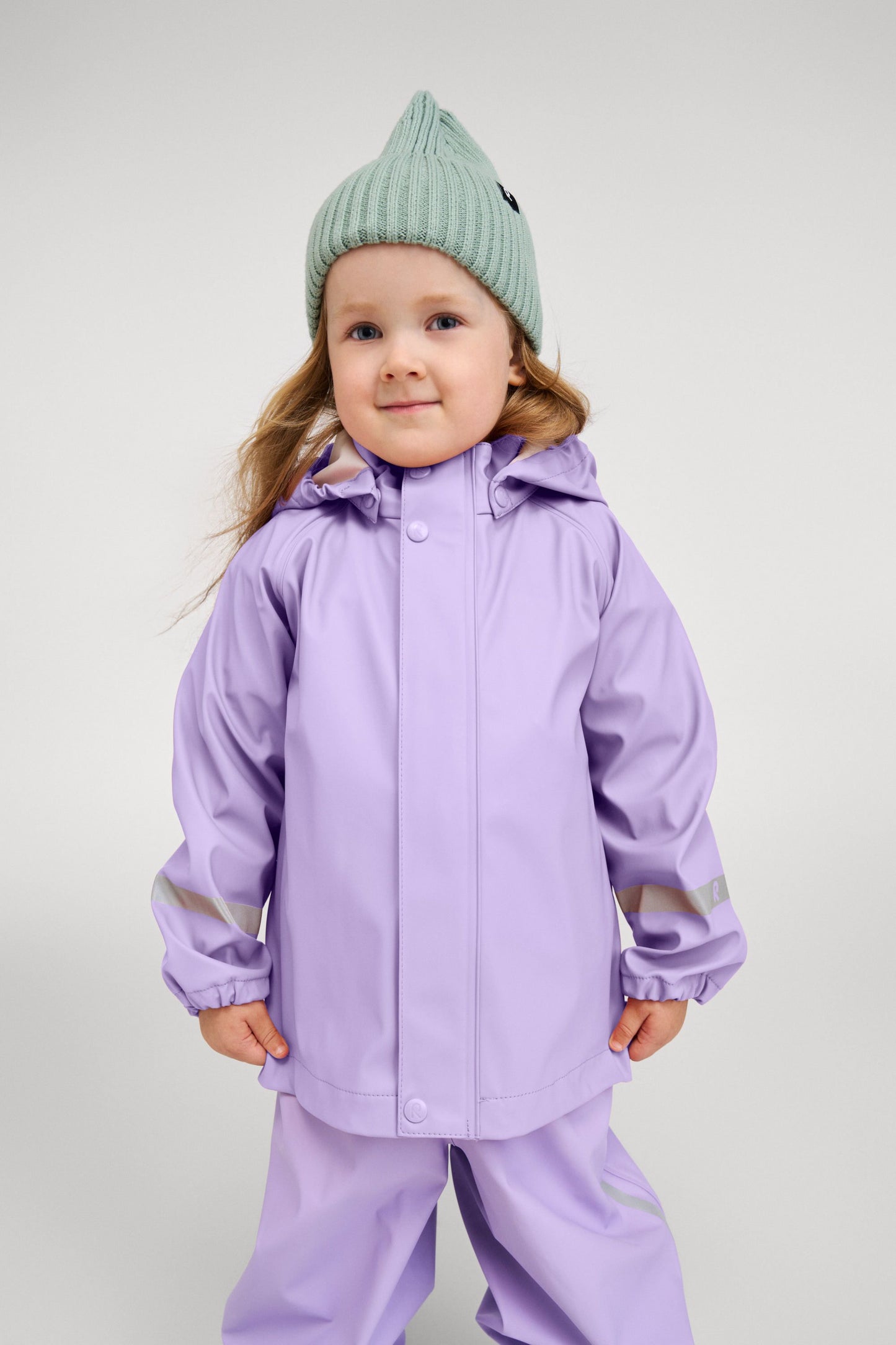 Lifestyle portrait of a young child happily wearing the Reima Lampi raincoat in blooming lilac, paired with matching rain pants and a mint green knit beanie, highlighting the jacket's comfortable, flexible fit designed for active outdoor play.