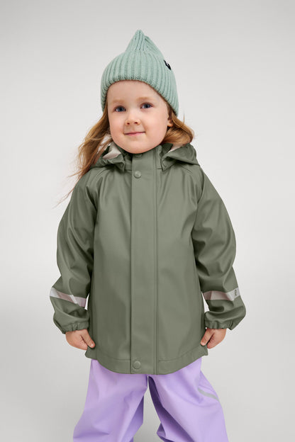 Lifestyle portrait of a smiling child comfortably wearing the greyish-green Reima Lampi raincoat paired with pastel purple rain pants and a knit beanie, illustrating the jacket's flexible, unrestrictive fit designed for active outdoor puddle jumping.