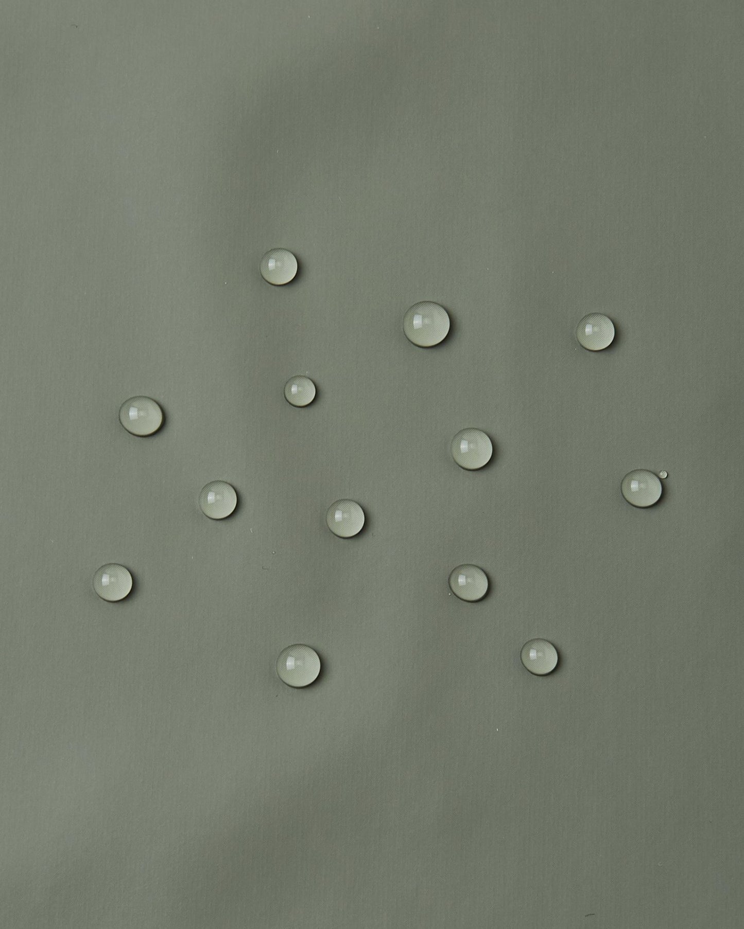 Close-up demonstration of water droplets tightly beading and rolling off the smooth waterproof polyurethane surface of the Reima Lampi greyish-green raincoat, proving its effectiveness at keeping kids completely dry during heavy downpours.