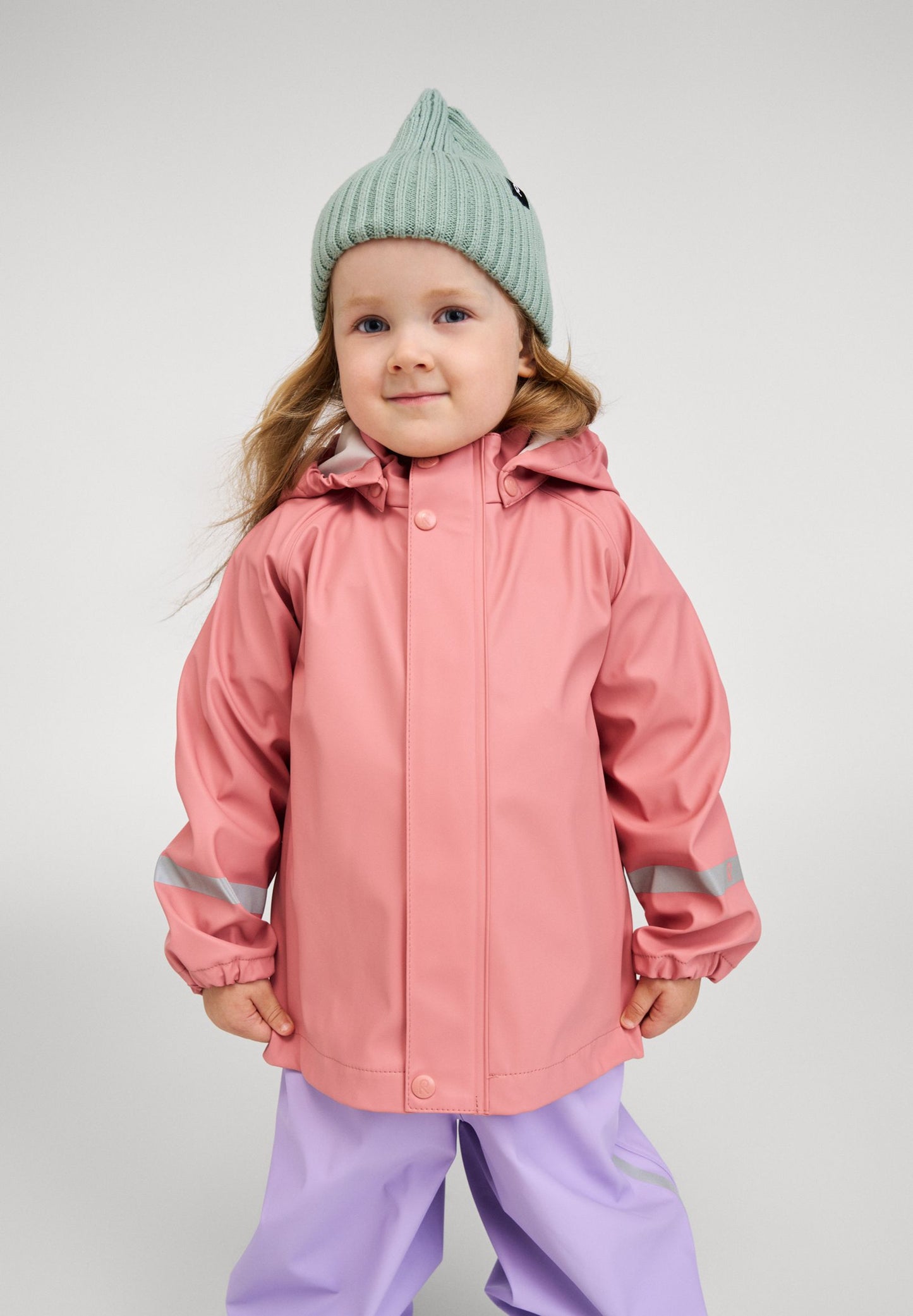 Lifestyle portrait of a young child wearing the Reima Lampi raincoat in rose blush, paired with light purple rain pants and a mint green knit beanie, showcasing the jacket's comfortable, unrestrictive fit designed for active rainy day puddle jumping.