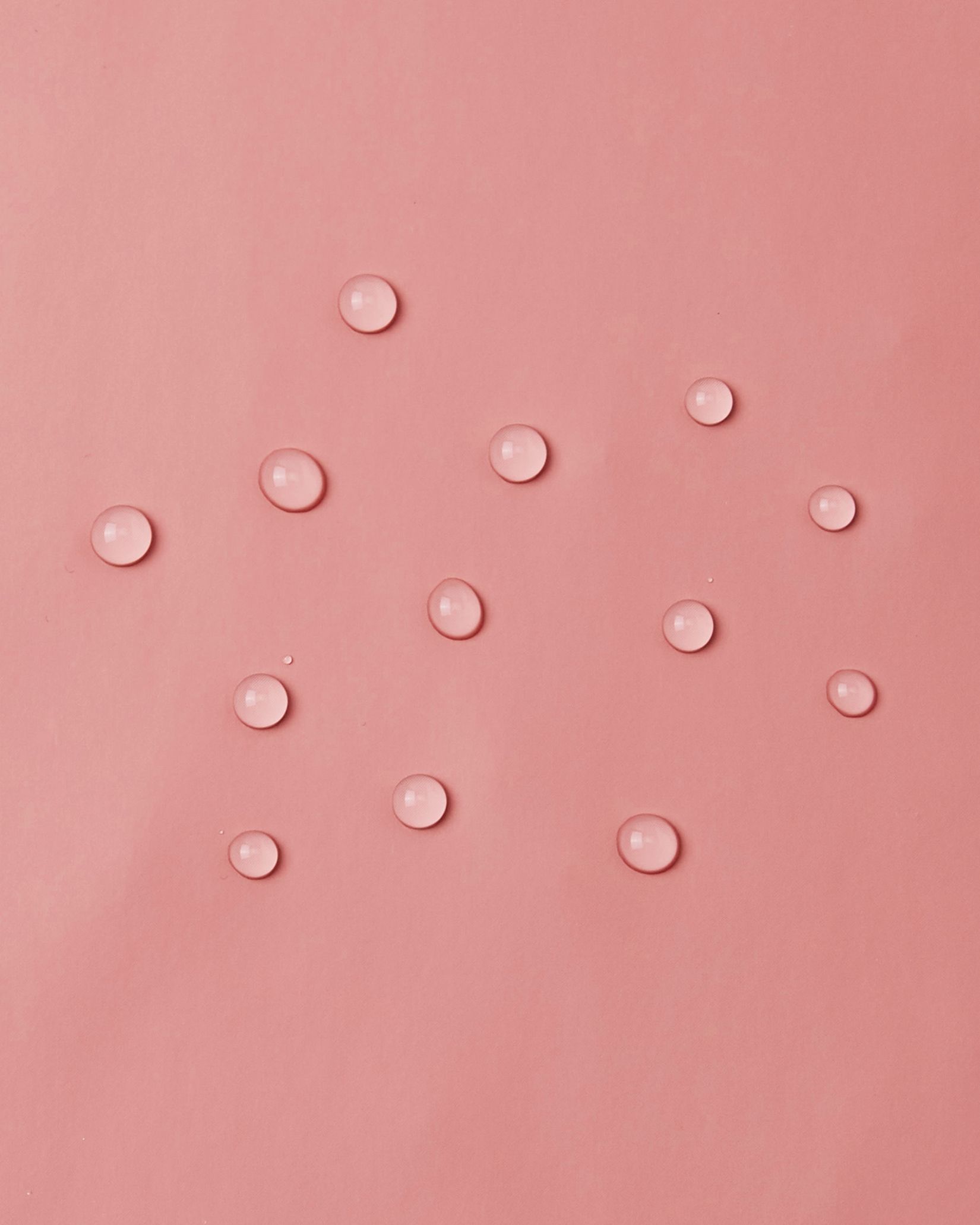 Macro close-up demonstrating water droplets perfectly beading and sitting lightly on top of the smooth waterproof polyurethane surface of the Reima Lampi rose blush raincoat, highlighting the material's excellent rain-repellent capabilities.