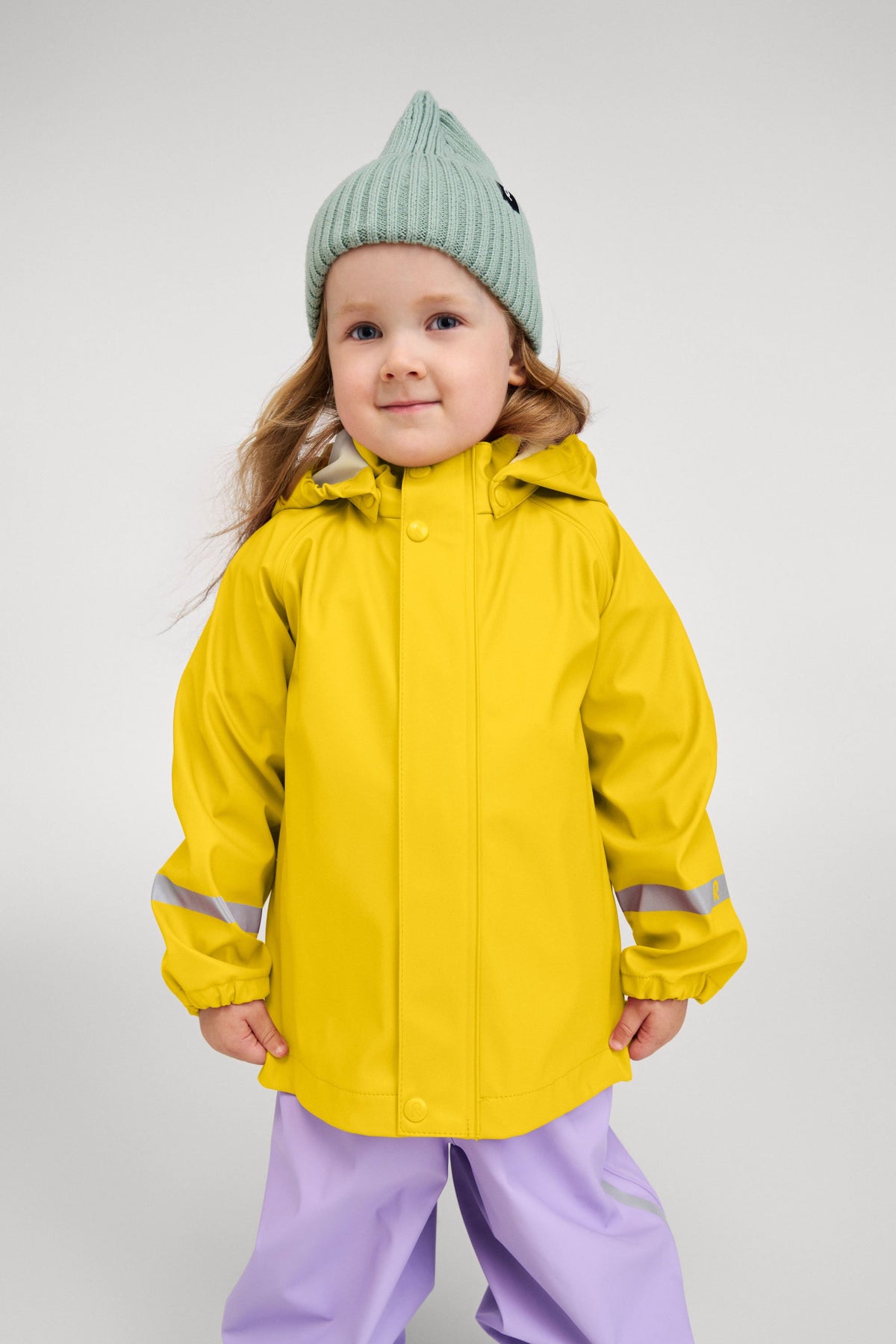 Lifestyle portrait of a young child wearing the bright yellow Reima Lampi raincoat paired with purple rain pants and a knit beanie, demonstrating a comfortable, flexible fit perfect for active outdoor play on rainy days.