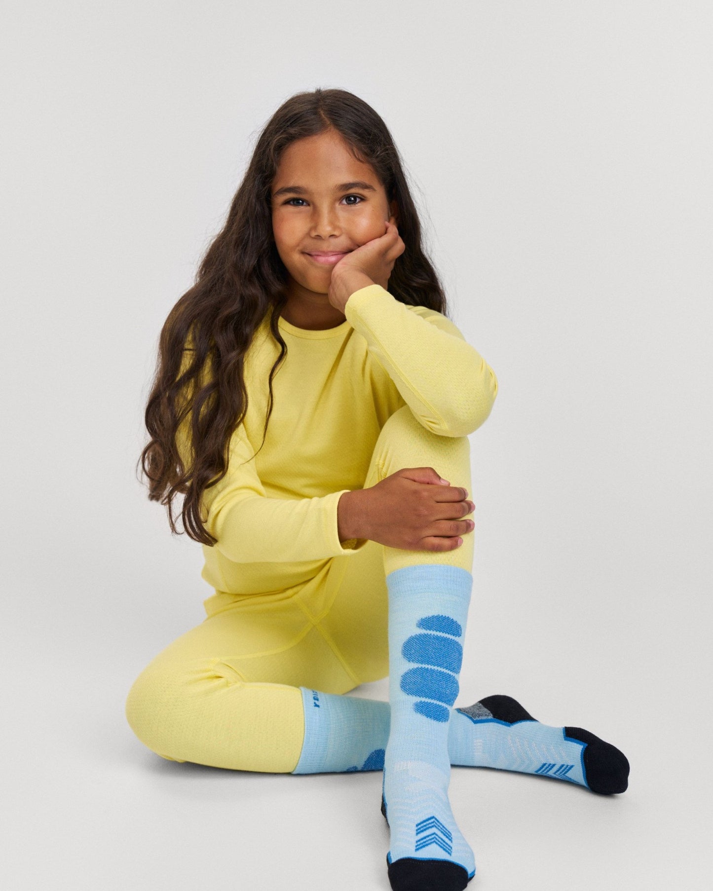 Lifestyle portrait of a smiling young girl sitting down and wearing the bright yellow Reima Lani kids' thermal base layer set. The image showcases the comfortable, temperature-regulating long-sleeve winter top and matching thermal underwear pants, paired with blue ski socks for cold weather layering.