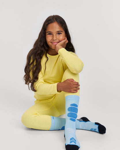 Lifestyle portrait of a smiling young girl sitting down and wearing the bright yellow Reima Lani kids' thermal base layer set. The image showcases the comfortable, temperature-regulating long-sleeve winter top and matching thermal underwear pants, paired with blue ski socks for cold weather layering.