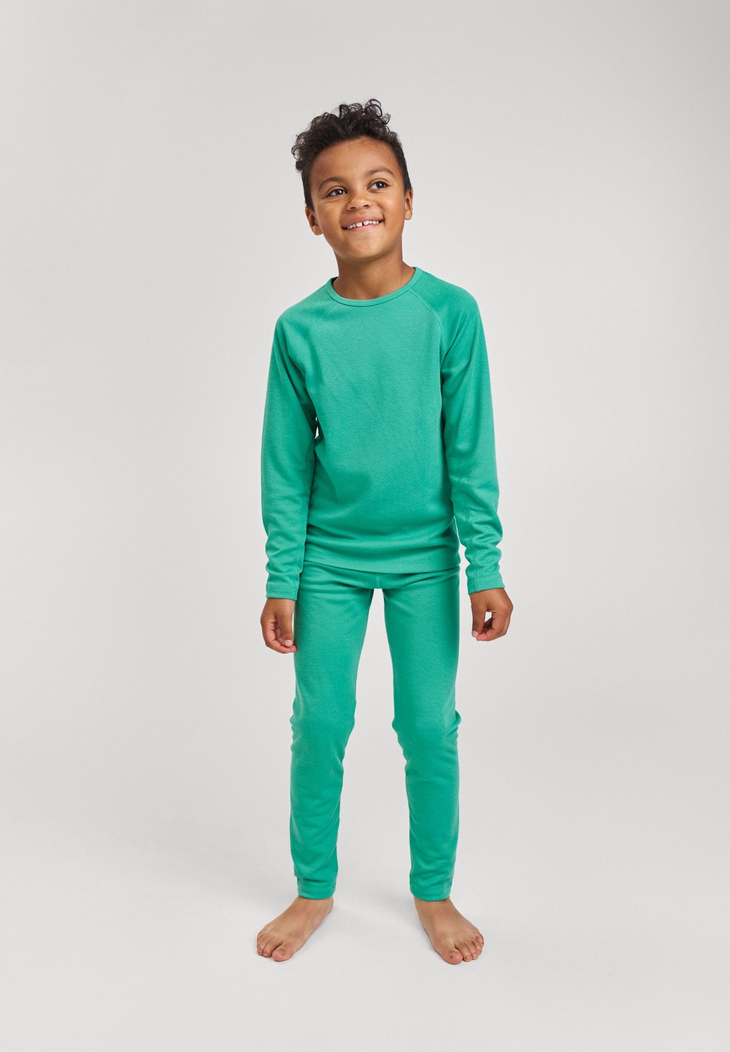 Lifestyle portrait of a young boy wearing the green Reima Lani kids' Thermolite base layer set, demonstrating its comfortable fit and flexibility for winter sports and daily wear.