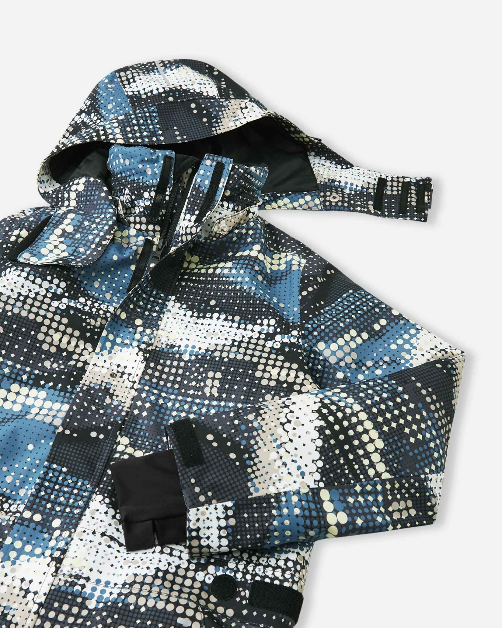 Upper front view of the Reima Lappea kids' winter ski suit showcasing the dynamic soft black, blue, and cream dot pattern, featuring a protective winter hood, a high wind-blocking collar, and a smooth, durable main zipper.