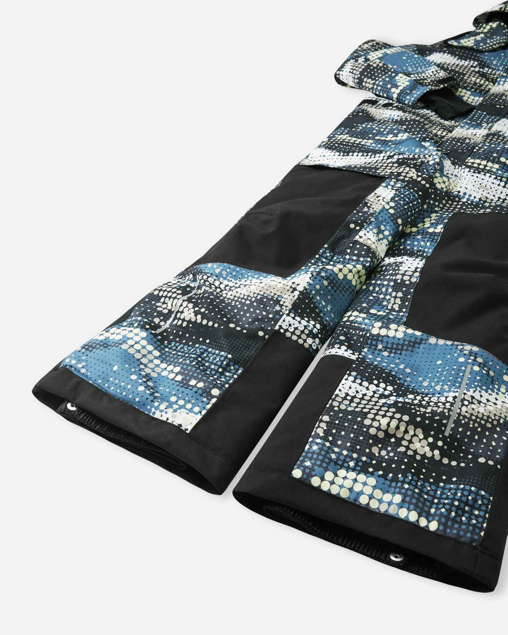 Close-up of the lower legs on the Reima Lappea kids' ski suit, showing the transition from solid black durable knee panels to patterned lower legs, complete with reinforced hems and adjustable snap button closures designed to fit securely over ski boots.