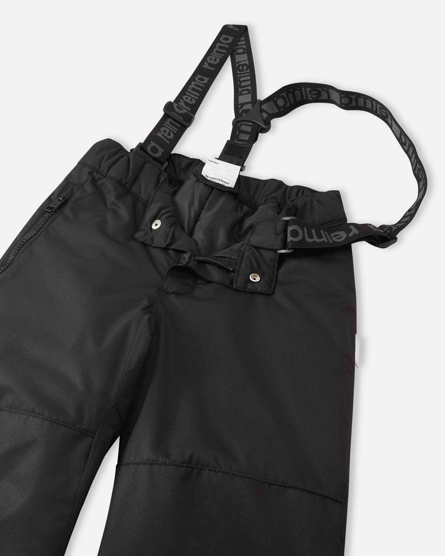 Close-up detail of the front waist on the black Reima Liikun kids' snow pants, showing the secure snap buttons, smooth zipper fly, elastic back waistband, and adjustable branded suspenders.