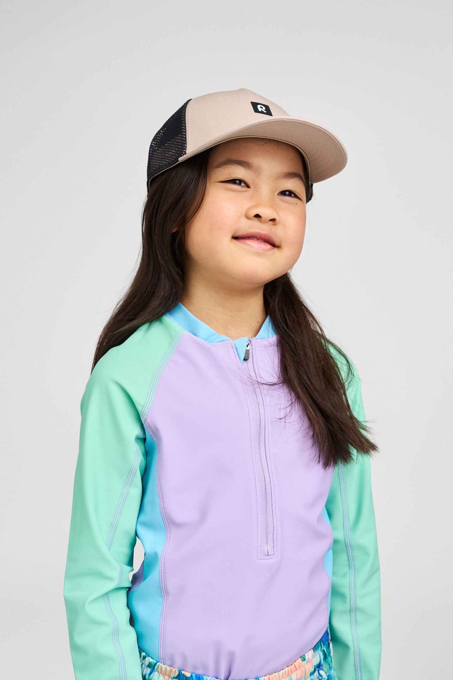 Lifestyle shot of a young girl wearing the Reima Liplatus kids' organic cotton trucker cap in earthy beige, demonstrating its comfortable fit and stylish, breathable design for active summer days.