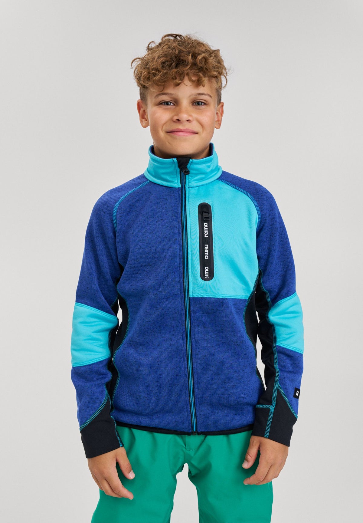 Lifestyle shot of a smiling boy wearing the twilight blue Reima Liukuen kids' fleece jacket paired with green outdoor pants, demonstrating the comfortable, active fit.