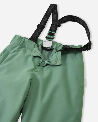 Close-up detail of the front waist on the green clay Reima Loikin kids' snow pants, showing the secure snap buttons, smooth zipper fly, elastic back waistband, and adjustable branded suspenders.