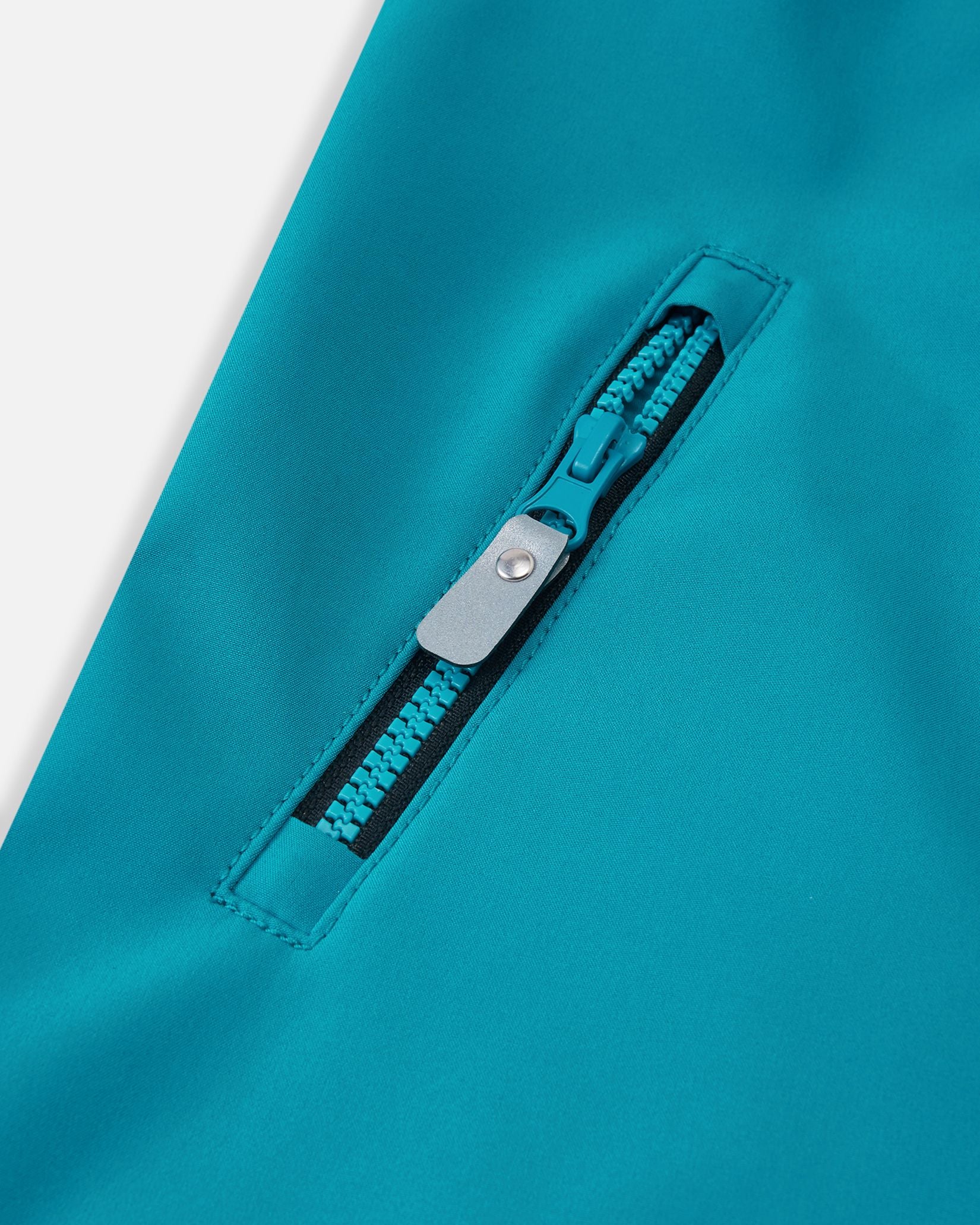 Close-up of the functional zippered chest pocket on the aquatic blue Reima Mjosa kids' softshell suit, highlighting the color-matched aquatic zipper and clean stitching.