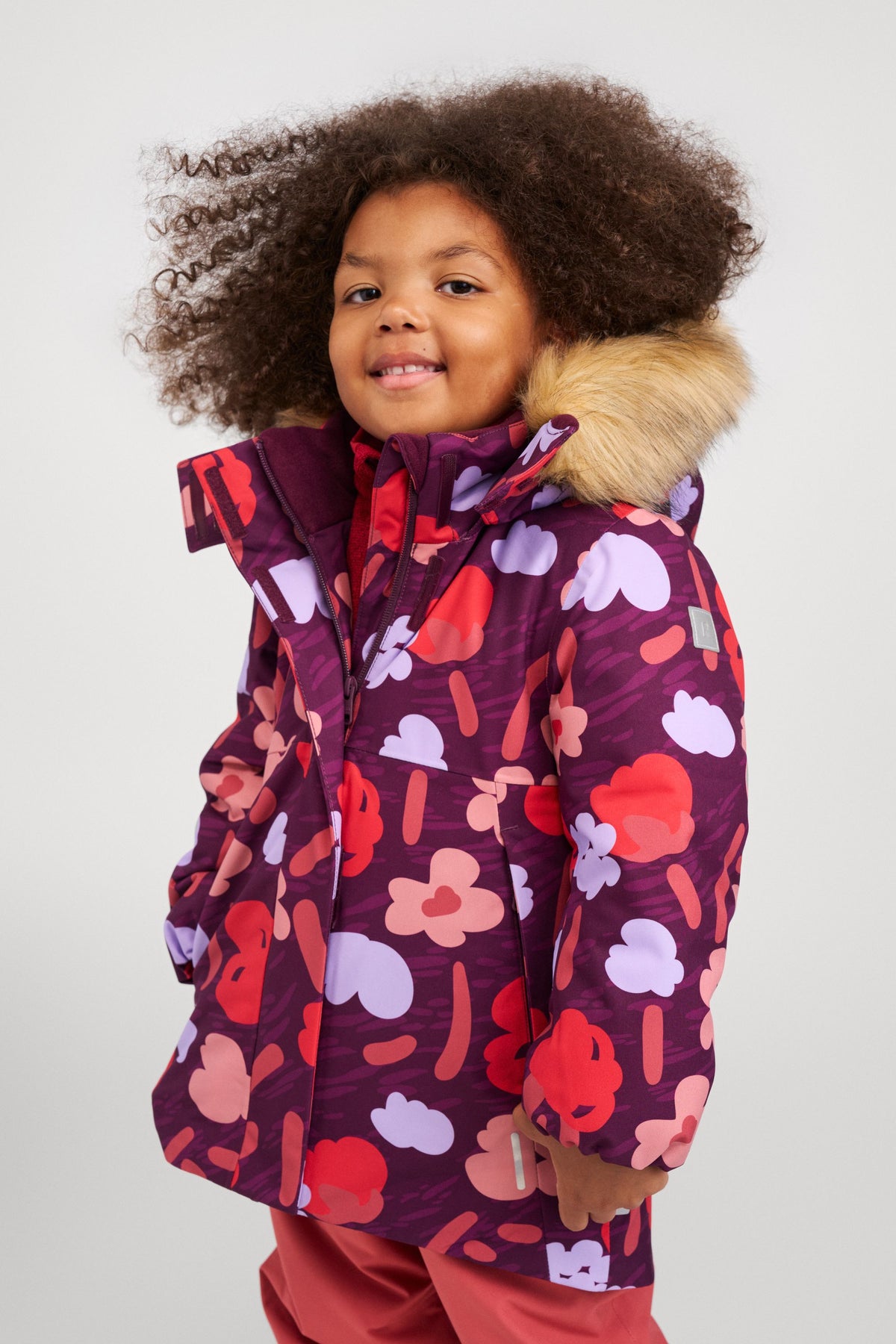 Lifestyle portrait of a young girl smiling and wearing the Reima Muurame girls' winter jacket in a vibrant deep purple, pink, and red abstract print. The image highlights the jacket's cozy faux fur-trimmed hood, high protective collar with a secure velcro wind flap, and longer parka-style fit, demonstrating its excellent warmth and weather protection for everyday winter wear.
