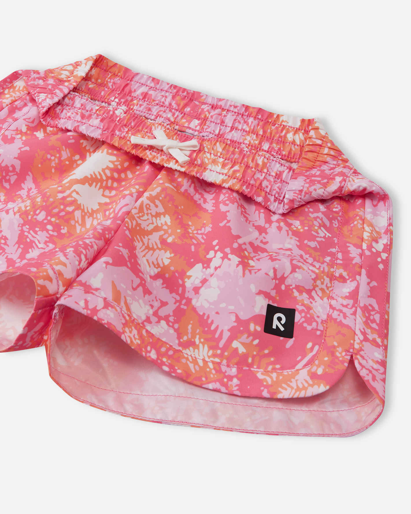 Close-up detail of the Reima Nauru girls' soft coral abstract swim shorts, focusing on the flexible gathered elastic waistband and the sturdy white inner drawstring that ensures the shorts stay secure.