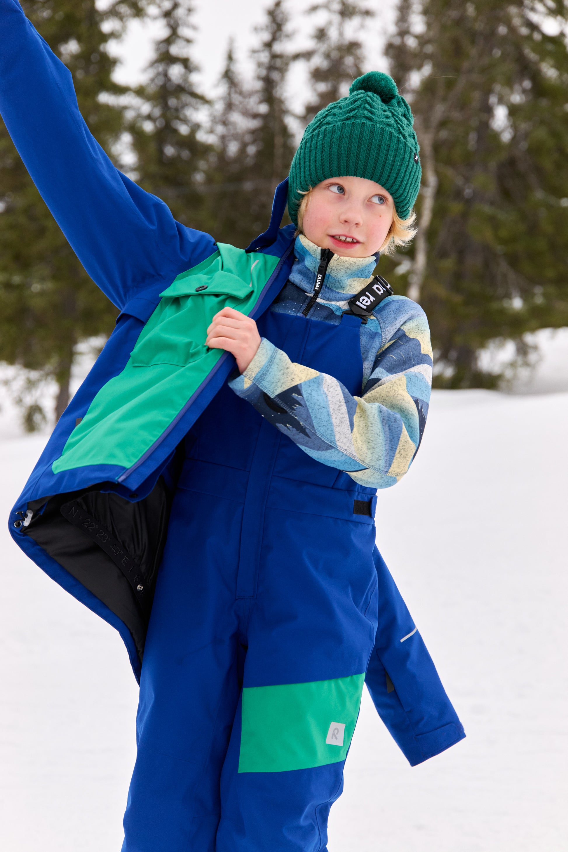 Lifestyle action shot of a child wearing the blue ocean Reima Niksini fleece mid-layer jacket under a ski jacket and overalls in a snowy mountain setting, demonstrating optimal cold-weather performance.