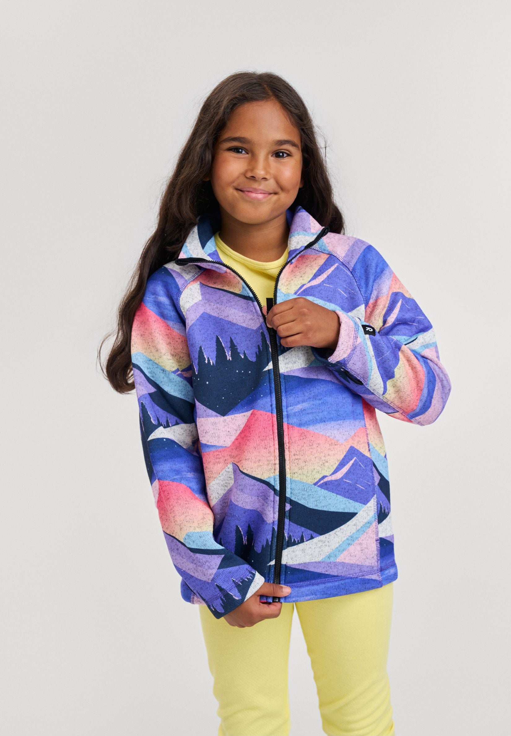 Lifestyle portrait of a young girl wearing the breezy violet Reima Niksini kids' fleece mid-layer jacket featuring a vibrant mountain graphic print, styled for active everyday outdoor wear.