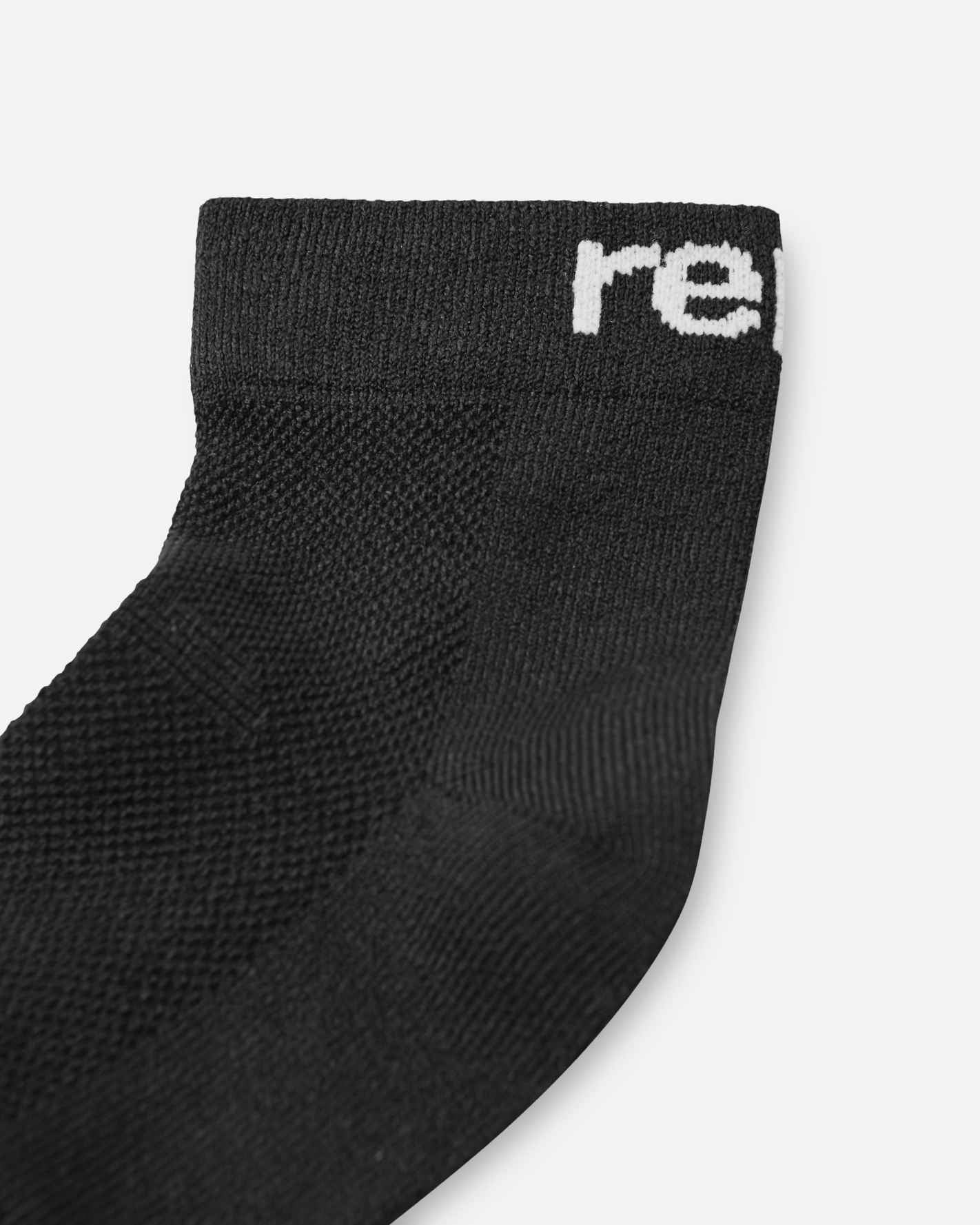 Macro close-up of the Reima Nilkat kids' black ankle sock, highlighting the breathable knit mesh fabric on the top of the foot and the contrasting white Reima logo woven into the ribbed cuff.