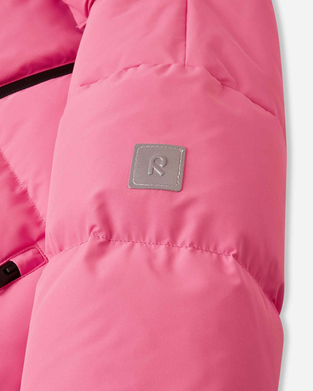 Close-up of the sleeve on the soft coral pink Reima Paimio children's puffer coat, highlighting the square reflective Reima 'R' logo patch that enhances visibility during dark winter days.
